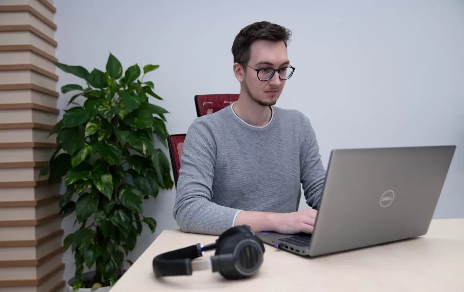 Java programátor Filip sedí v kancelárií na stoličke pri stole kde je položený notebook a bezdrôtové slúchadlá