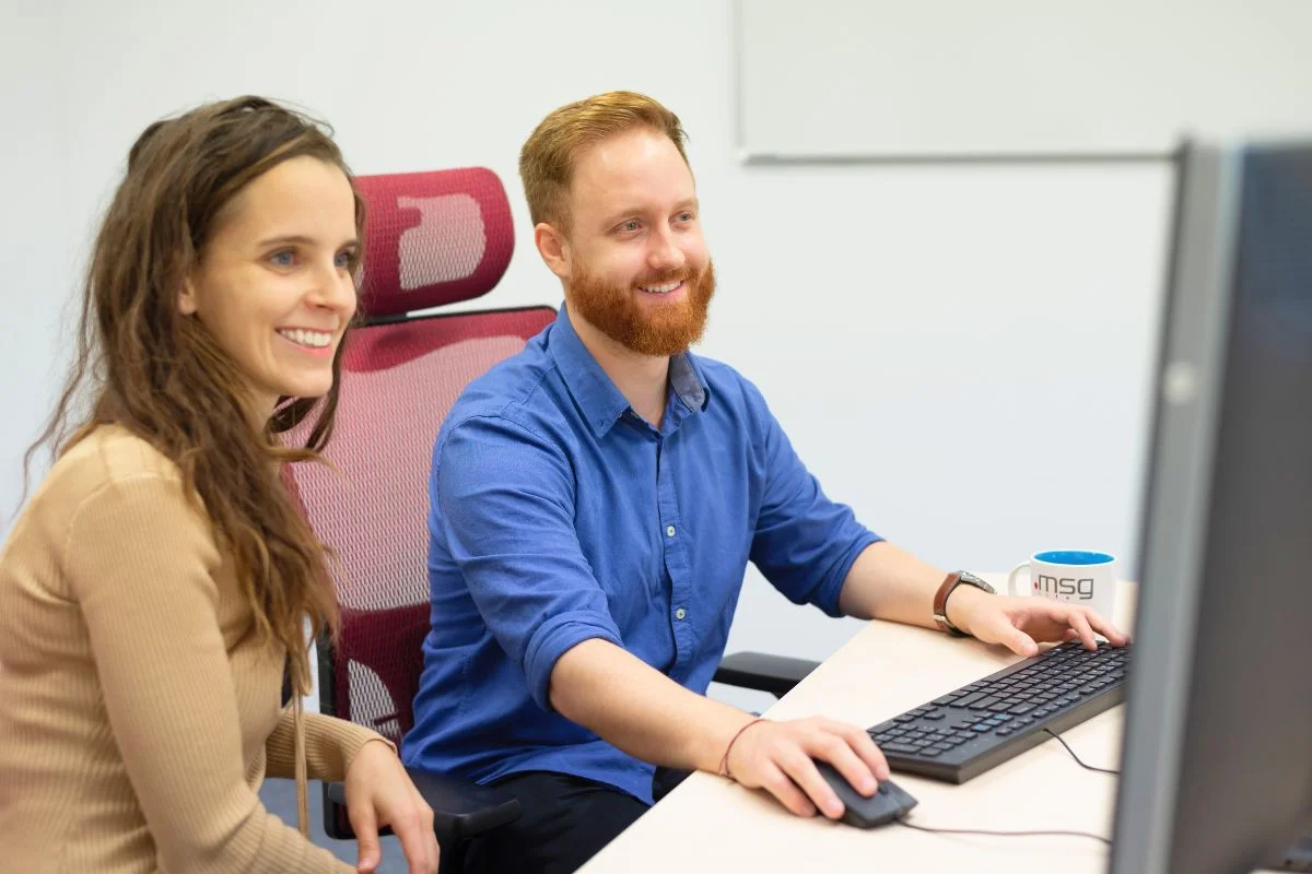spolupraca-napriec-timami-msg – msg life Slovakia Mladá žena a muž s ryšavou bradou sedia pri jednom počítači a usmievajú sa počas práce.